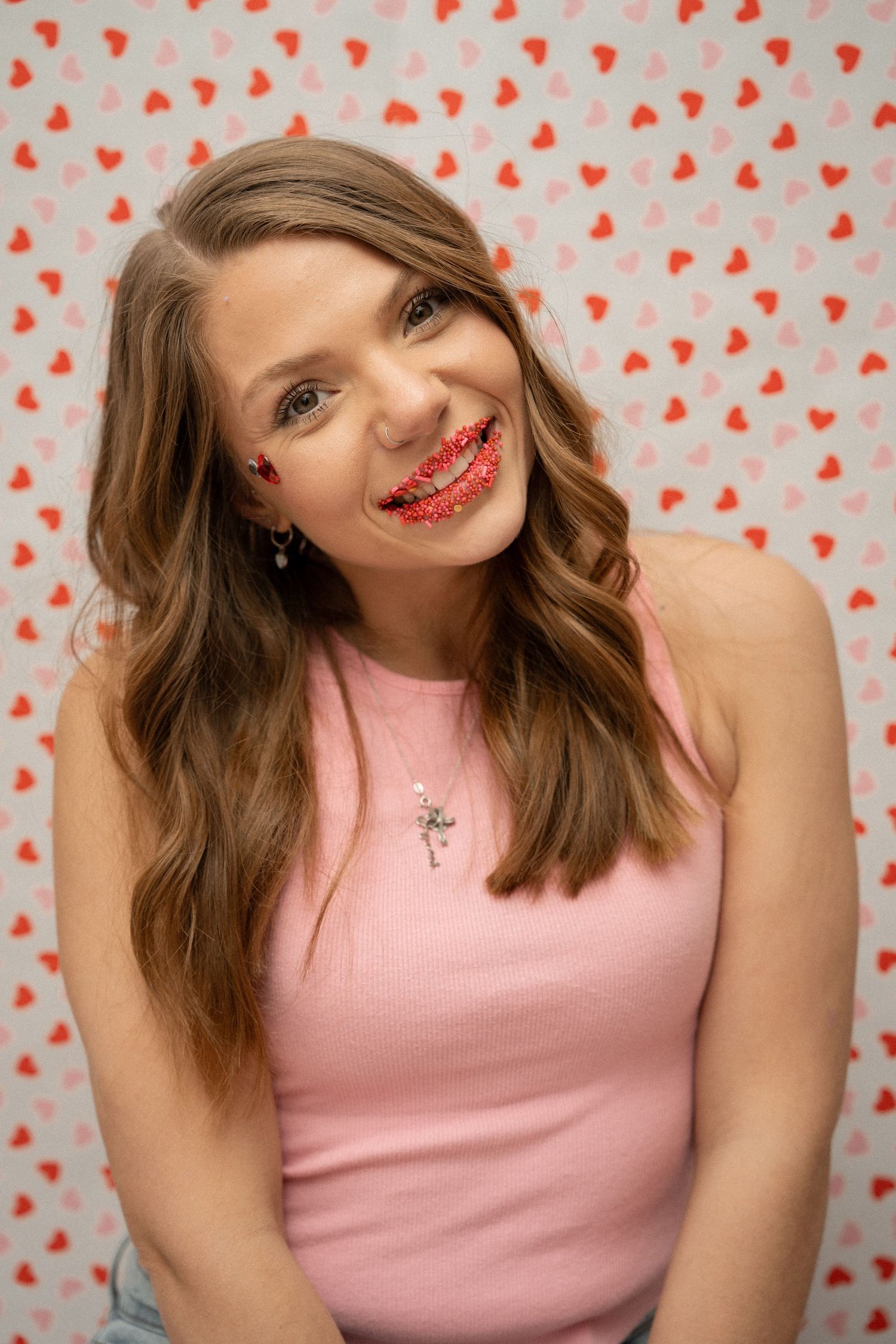 Smiling model in pink sleeveless top poses against heart-patterned background with glittery red lipstick.
