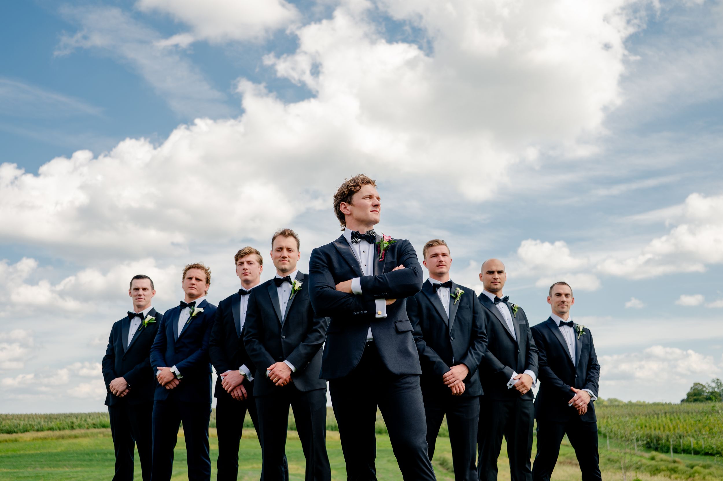 Groom in navy tuxedo stands confidently with groomsmen in matching suits outdoors under a blue sky.