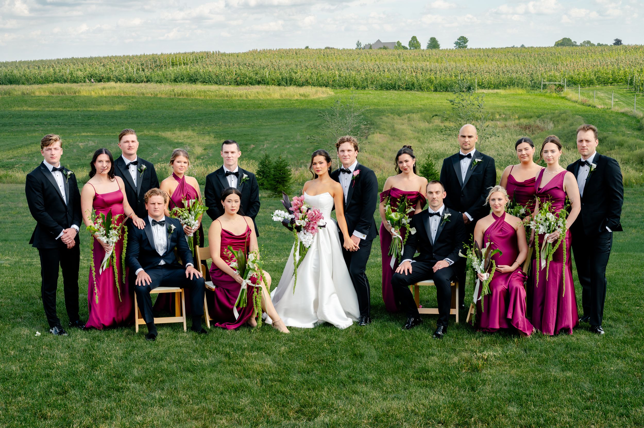 Wedding party in black tuxedos and burgundy gowns posing on green lawn with cornfield backdrop.