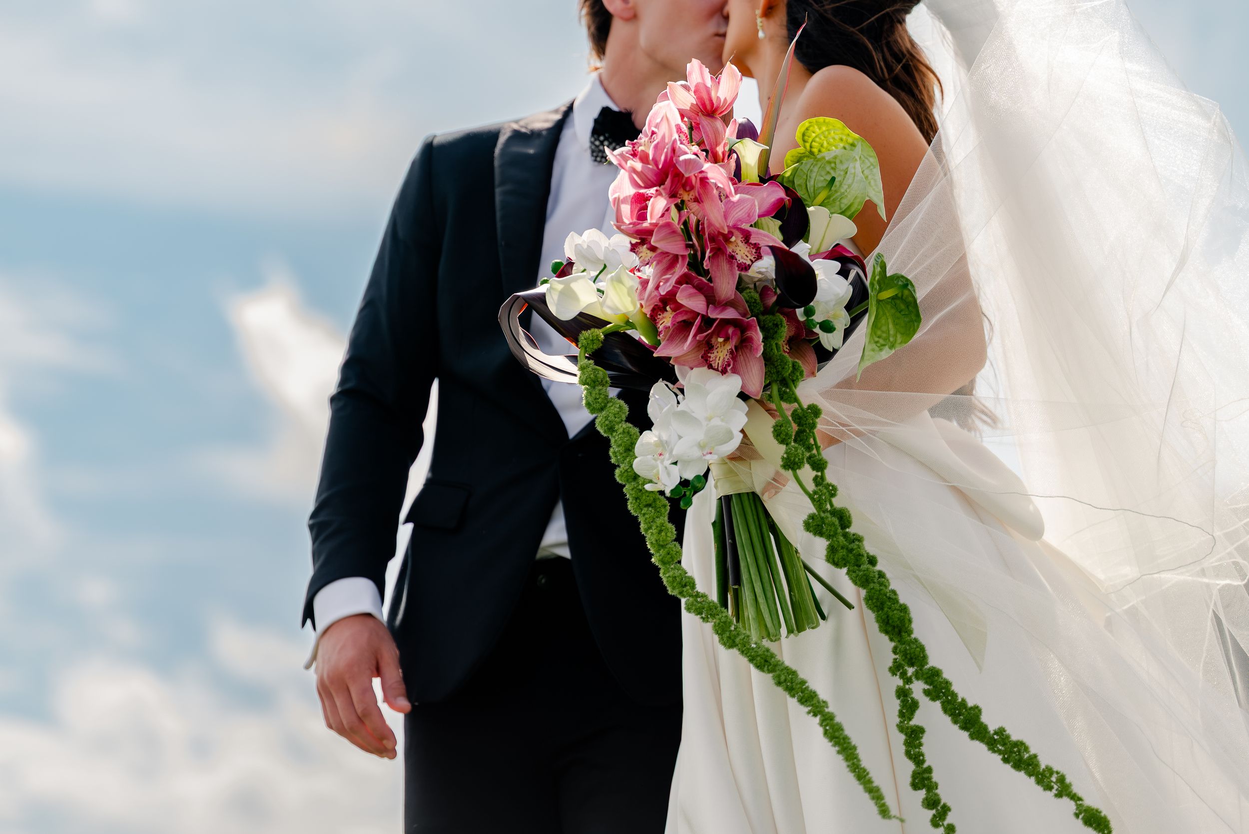 A wedding couple kissing, bride holding a pink and white floral bouquet with green trailing vines, groom in black tuxedo.