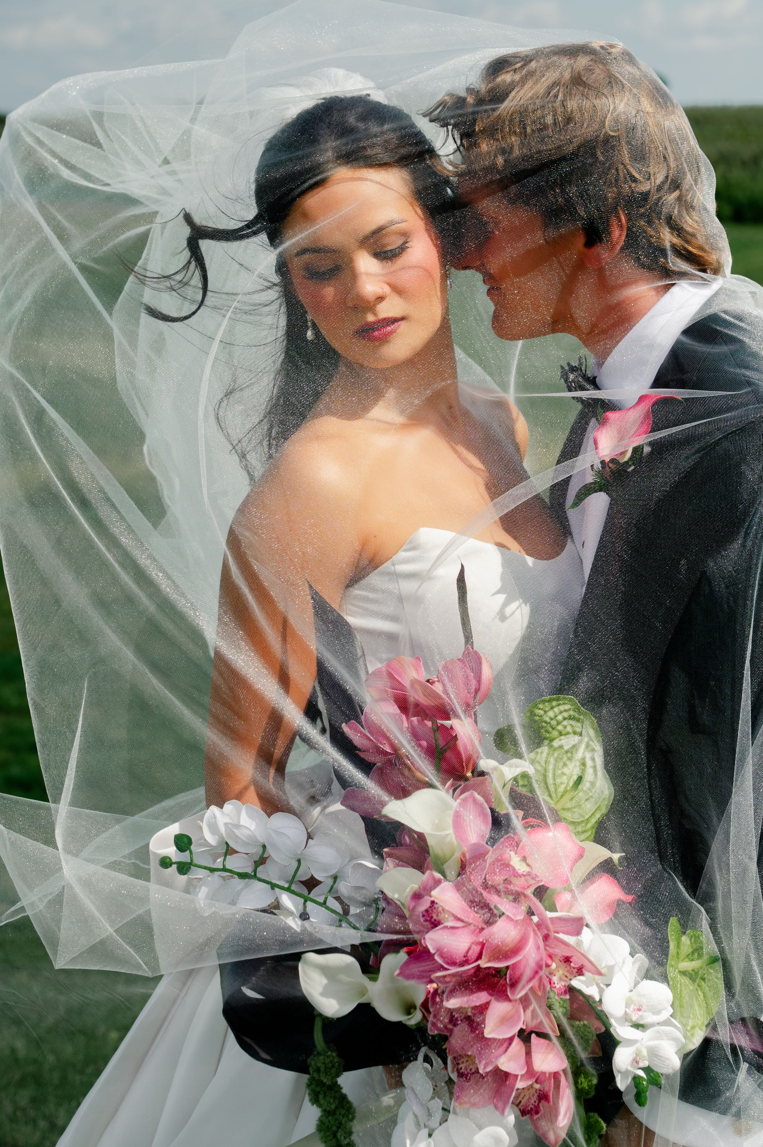 Bride and groom share romantic moment under flowing wedding veil, bride holding pink and white floral bouquet outdoors.