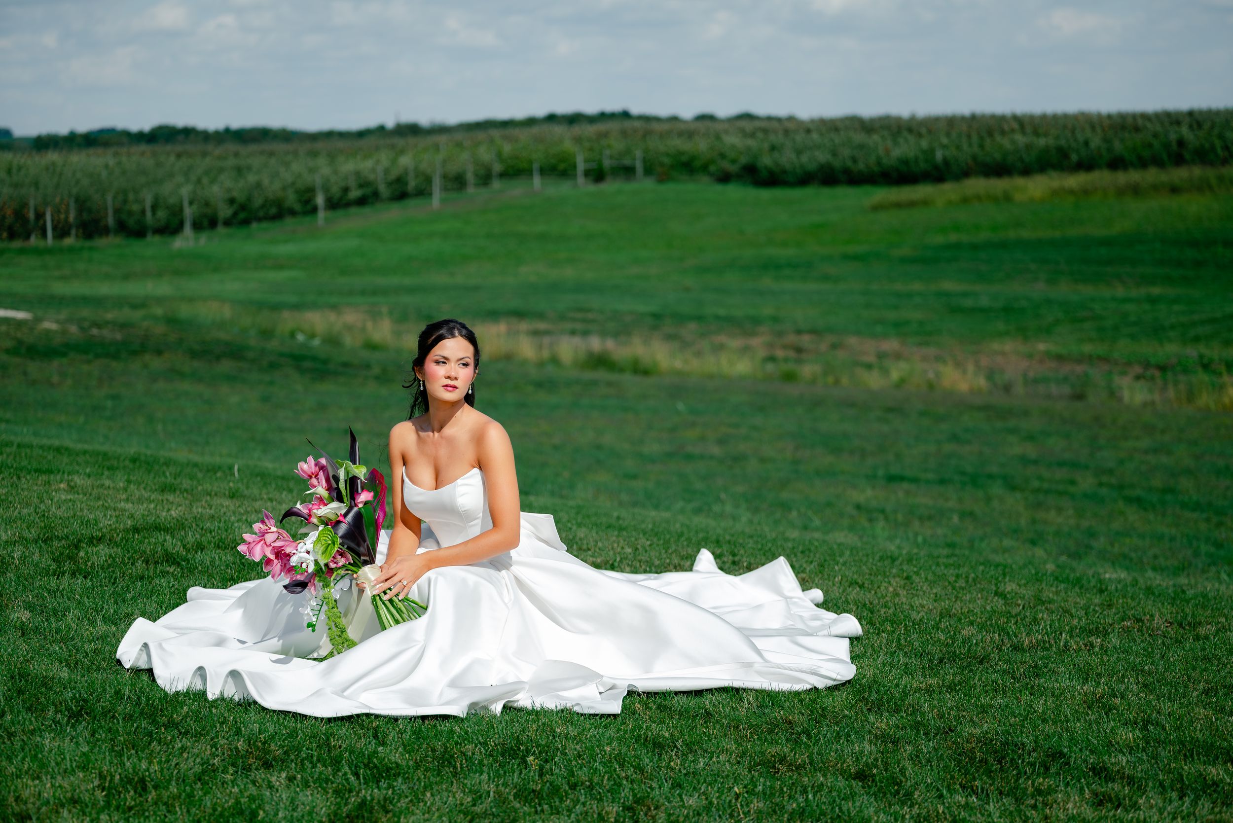 Bride in white strapless gown sitting on lush green grass with pink floral bouquet at outdoor wedding venue.