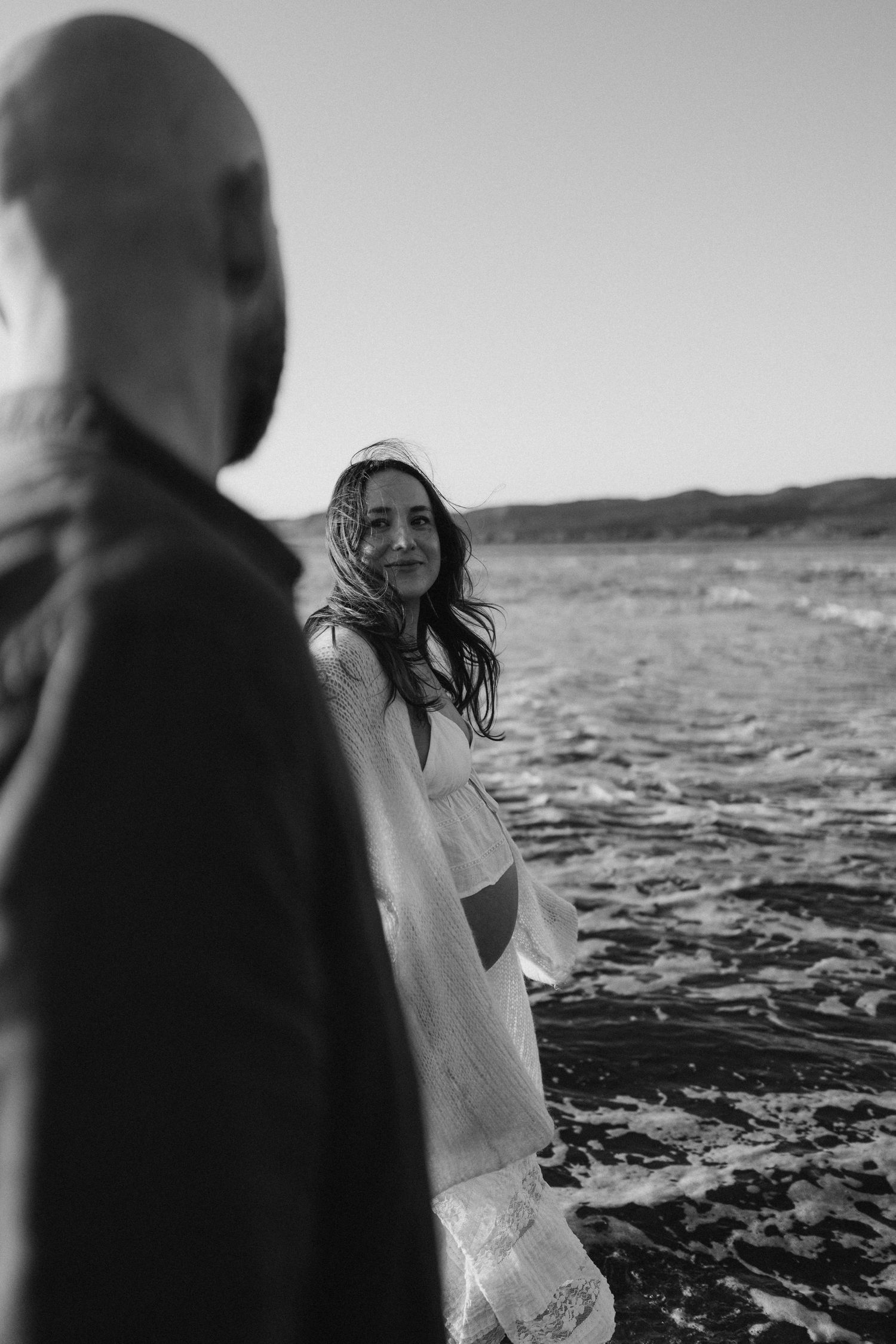 Black and white beach photo of a smiling woman in white dress with wind-blown hair, standing in ocean waves.