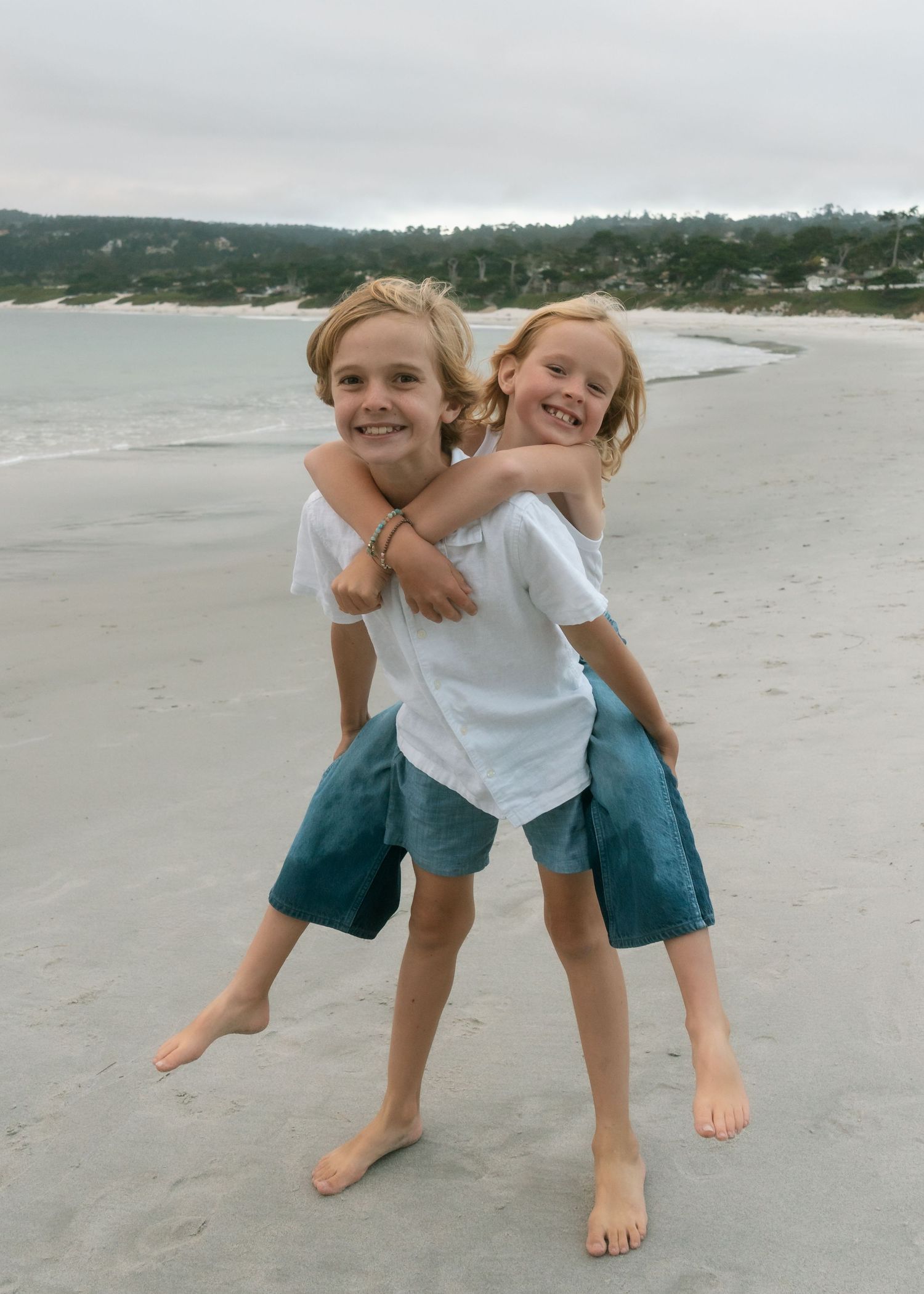 Two smiling children in white shirts play piggyback on a sandy beach with ocean waves and trees in the background.