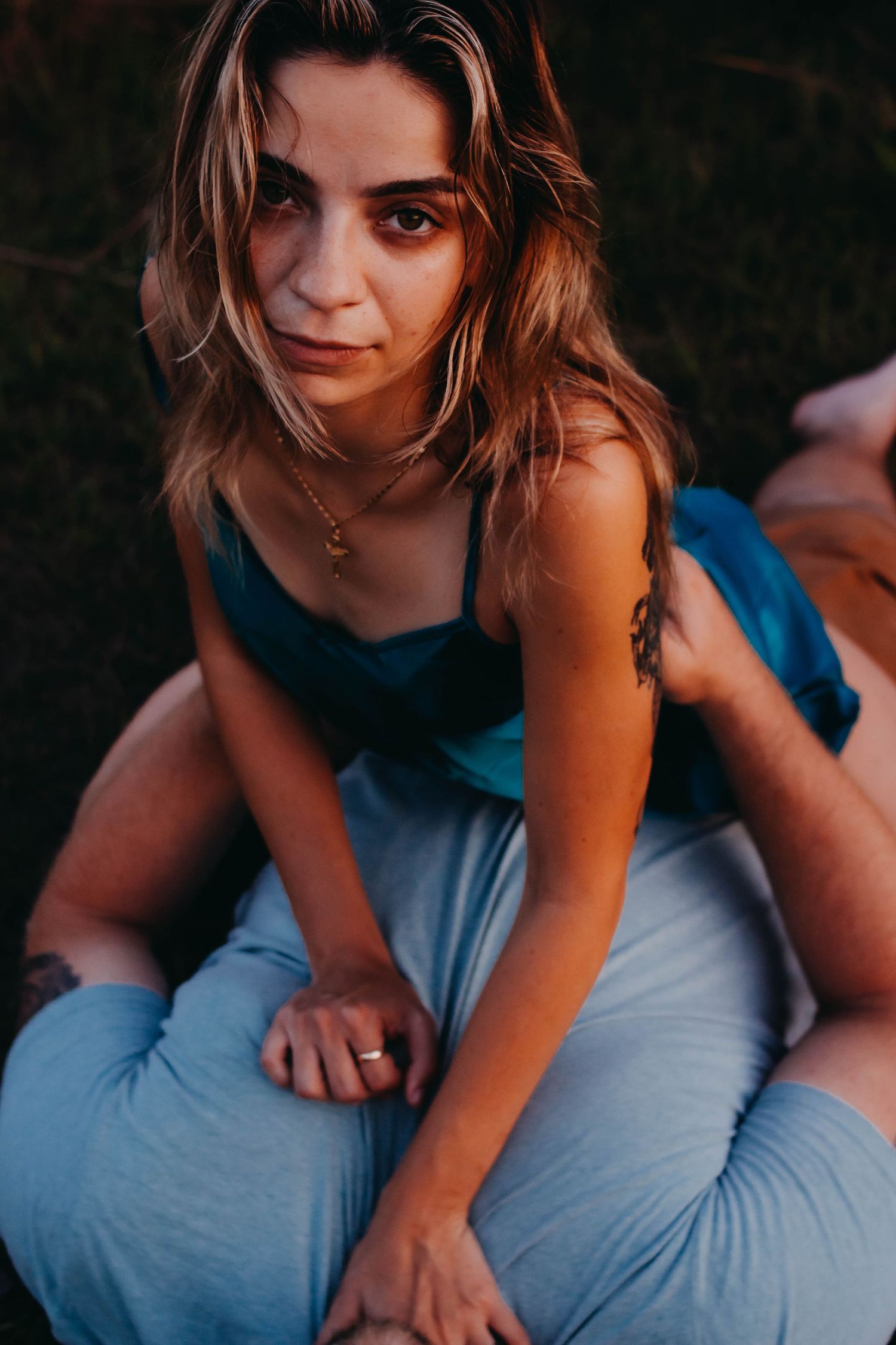 Young woman in blue dress sitting outdoors, wearing gold necklace and ring, with tattoos, looking at camera.