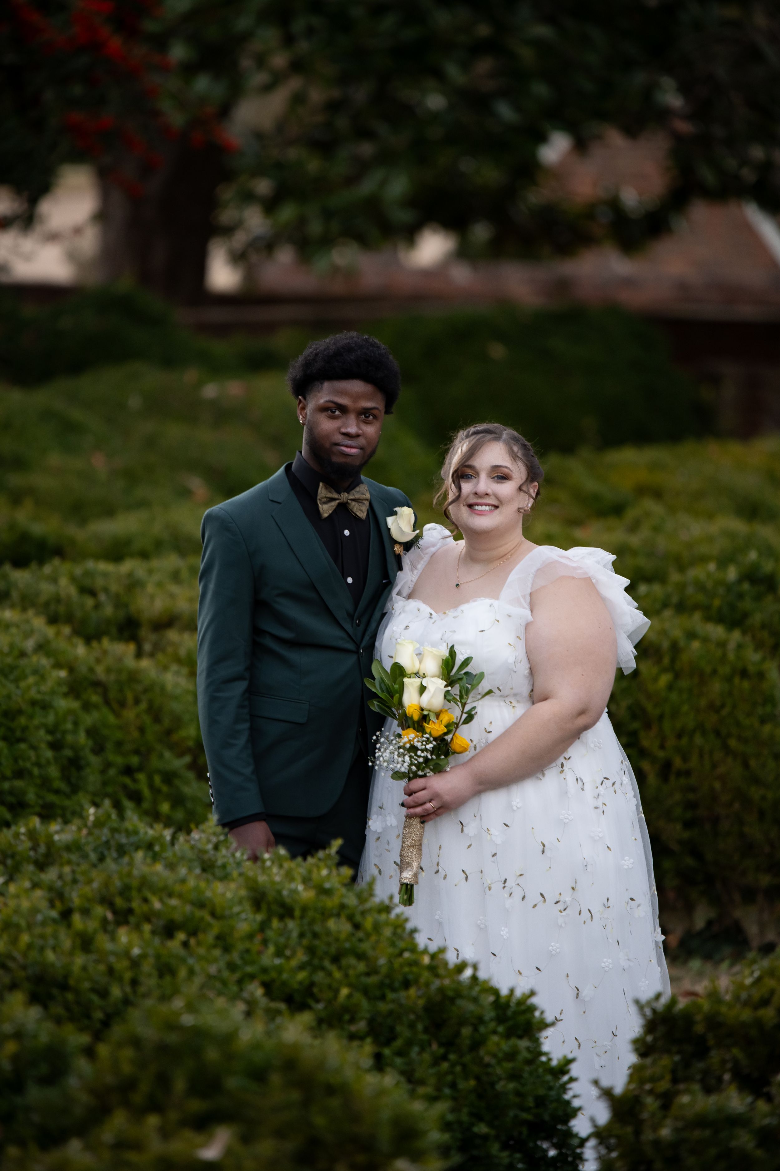 A couple in wedding attire pose together in a garden setting with green hedges and yellow flowers.