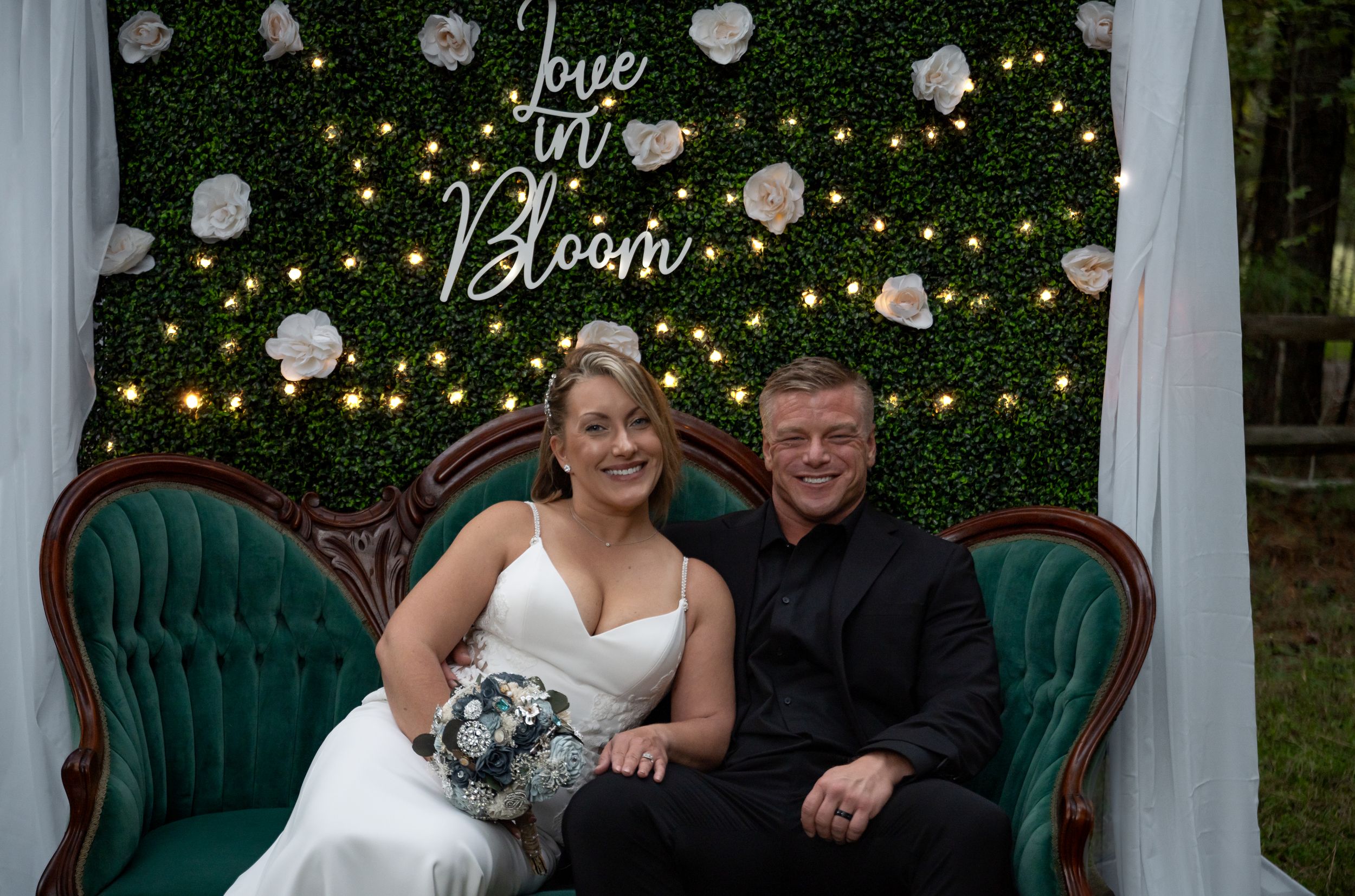 Wedding couple sits on emerald velvet couch against floral wall with string lights and 'Love in Bloom' sign.