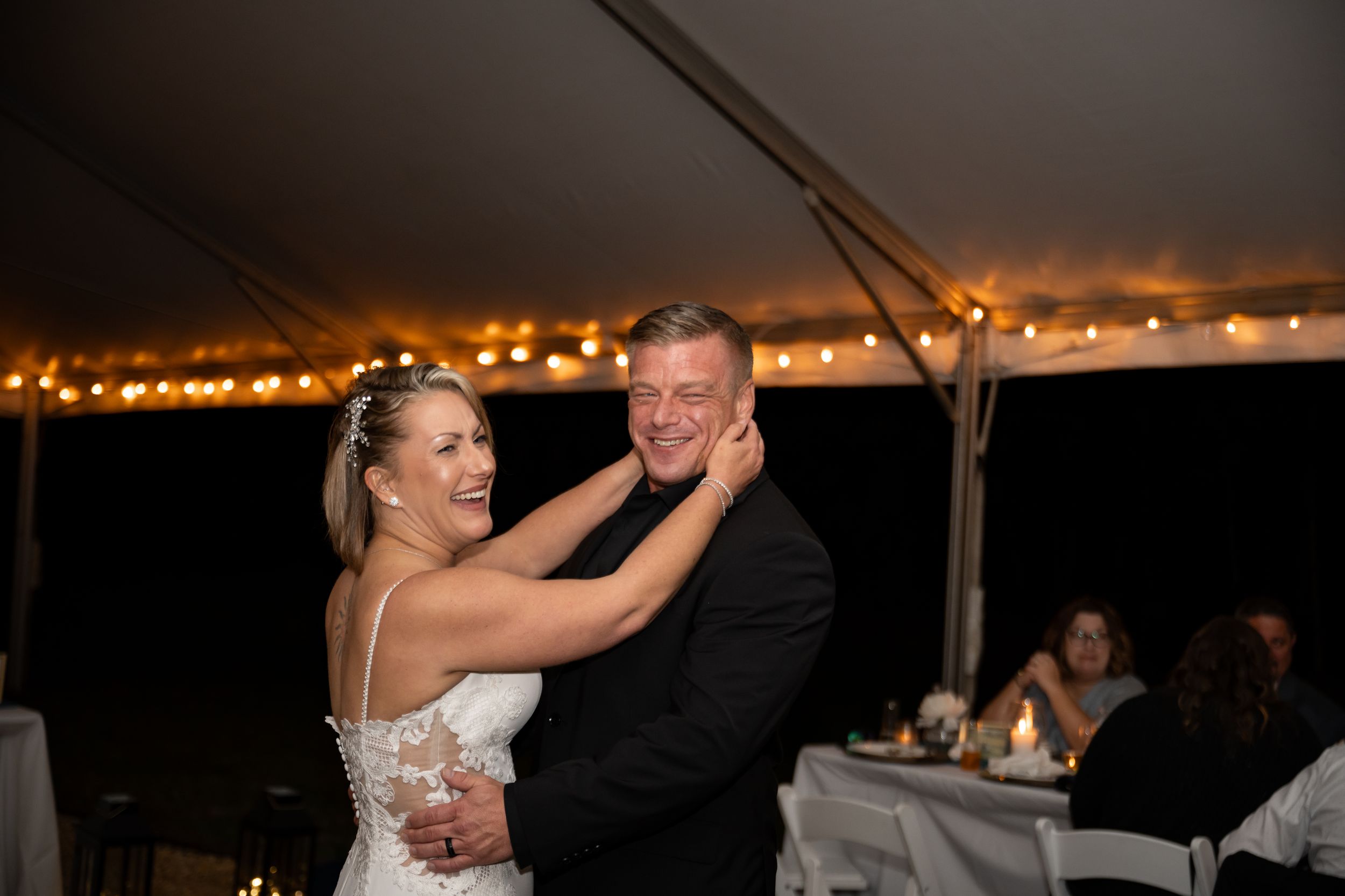 Dancing couple embraces during evening wedding reception under white tent with lights.