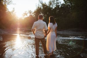 Couple stands together on rocky riverbank during golden hour sunset.