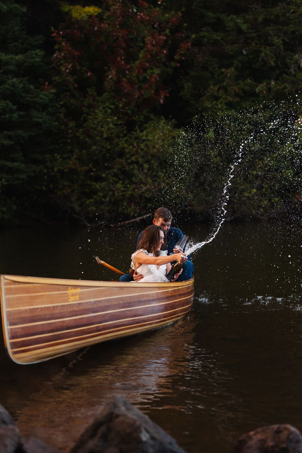 A couple shares a romantic kiss in a wooden canoe as water splashes around them.