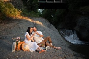 Couple enjoys picnic on riverbank beneath bridge at sunset.