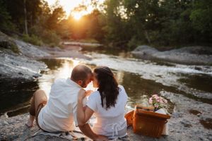 Couple sits together on rocky riverbank with picnic basket at golden hour.