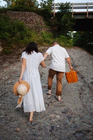A couple walking away on rocky terrain carrying a picnic basket and hat.
