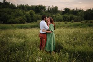 A romantic couple embraces in a lush green meadow at sunset wearing contrasting red pants and mint dress.