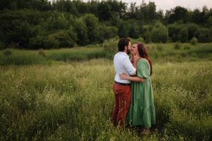 Couple shares a tender moment in a green summer meadow with forest backdrop.
