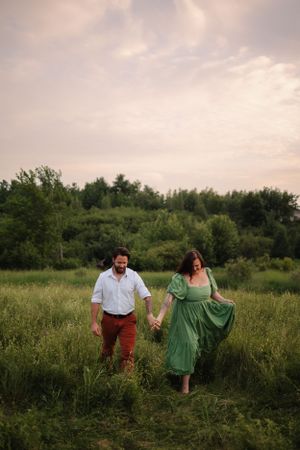 Couple walking hand in hand through tall grass in a meadow at dusk.