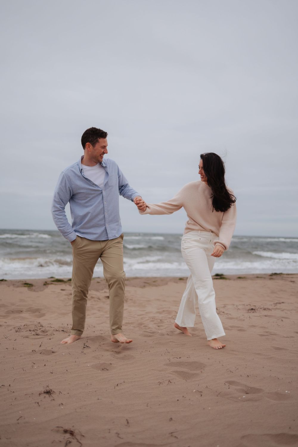 A playful couple runs along a sandy beach wearing light colored clothing.