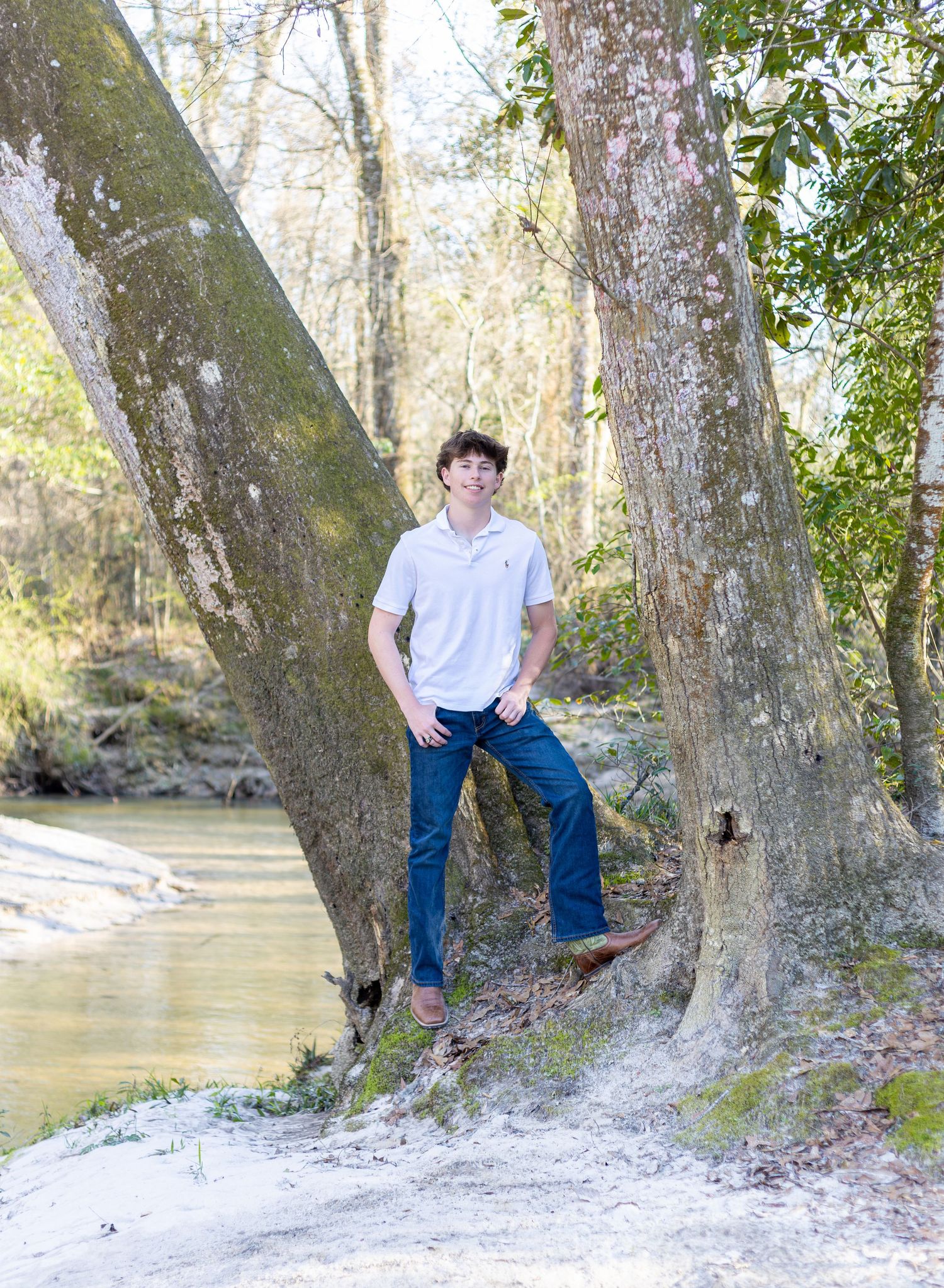 A photo that shows senior boy leaning against a large tree trunk beside a peaceful river on a sunny day.