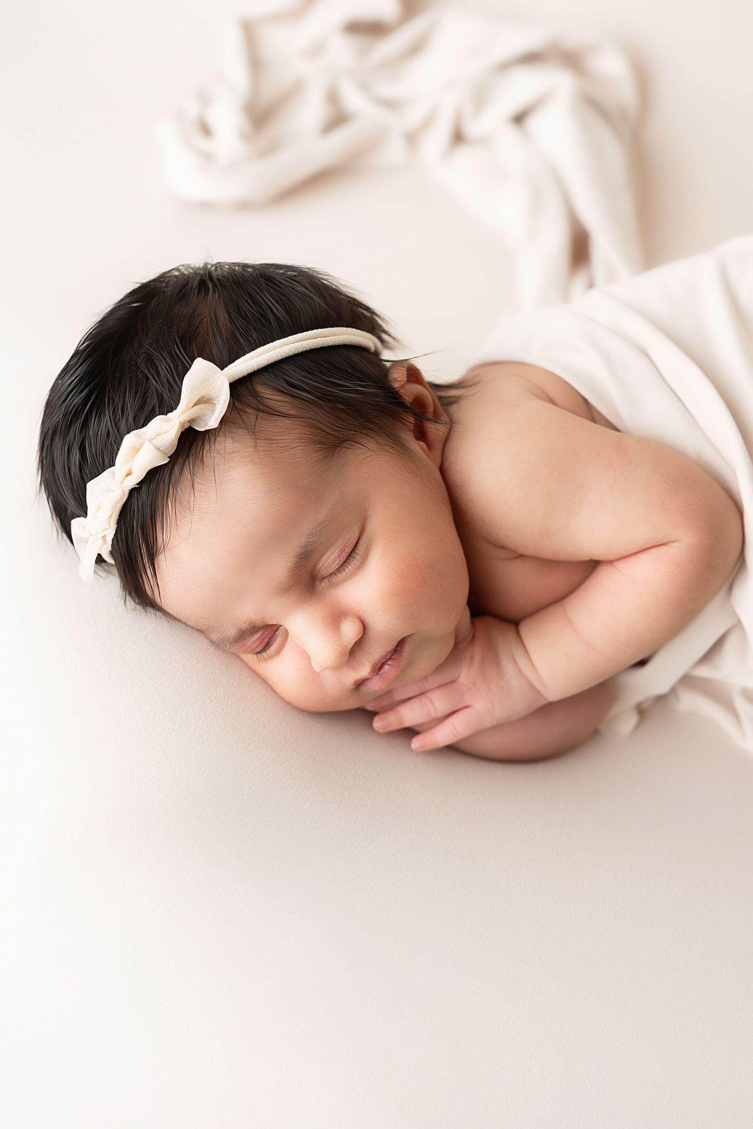 Peaceful newborn wearing white headband resting on soft white background.