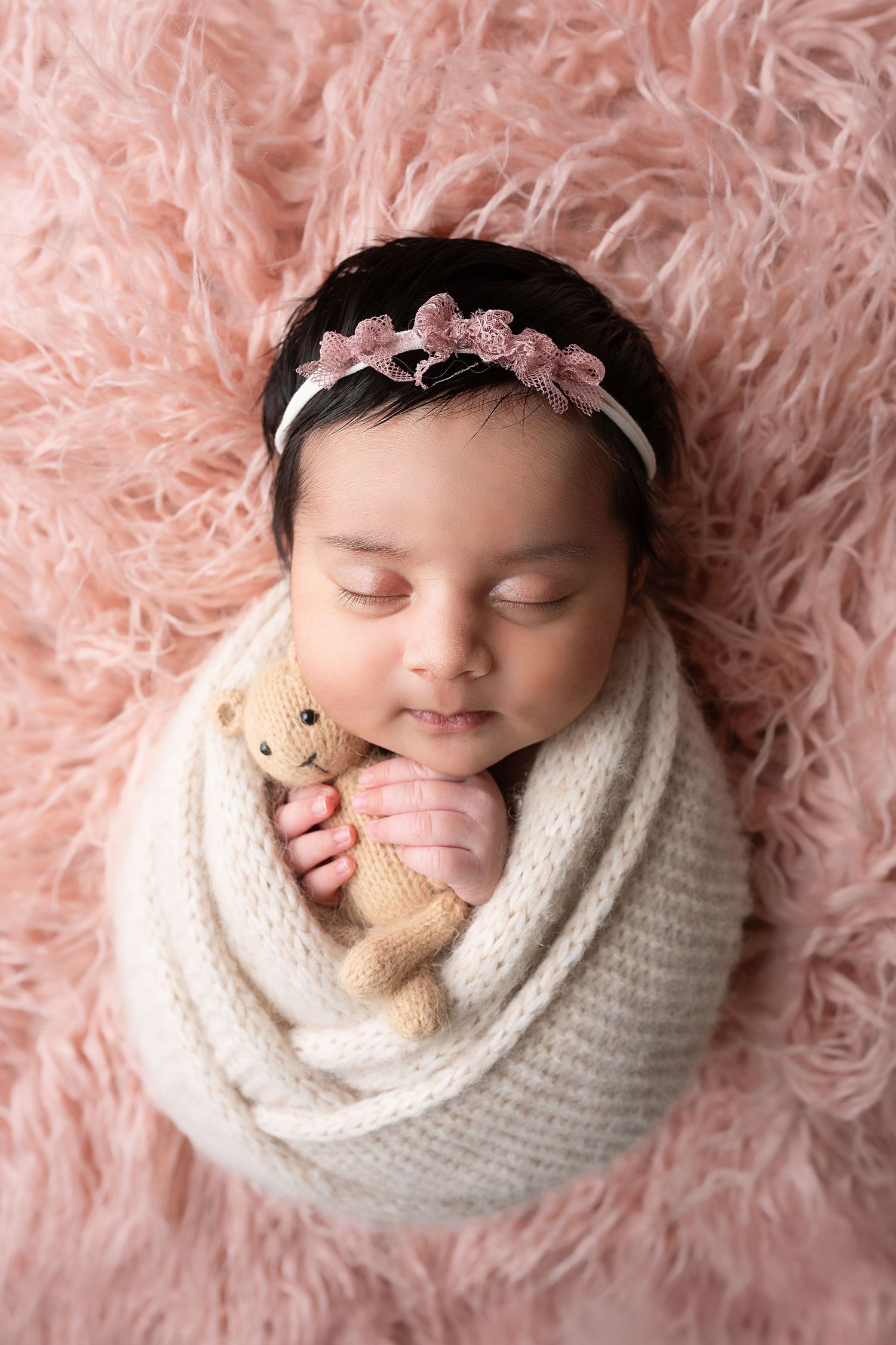 Newborn baby wrapped in white knit wrap with pink headband sleeping on pink fur backdrop holding tiny teddy bear.