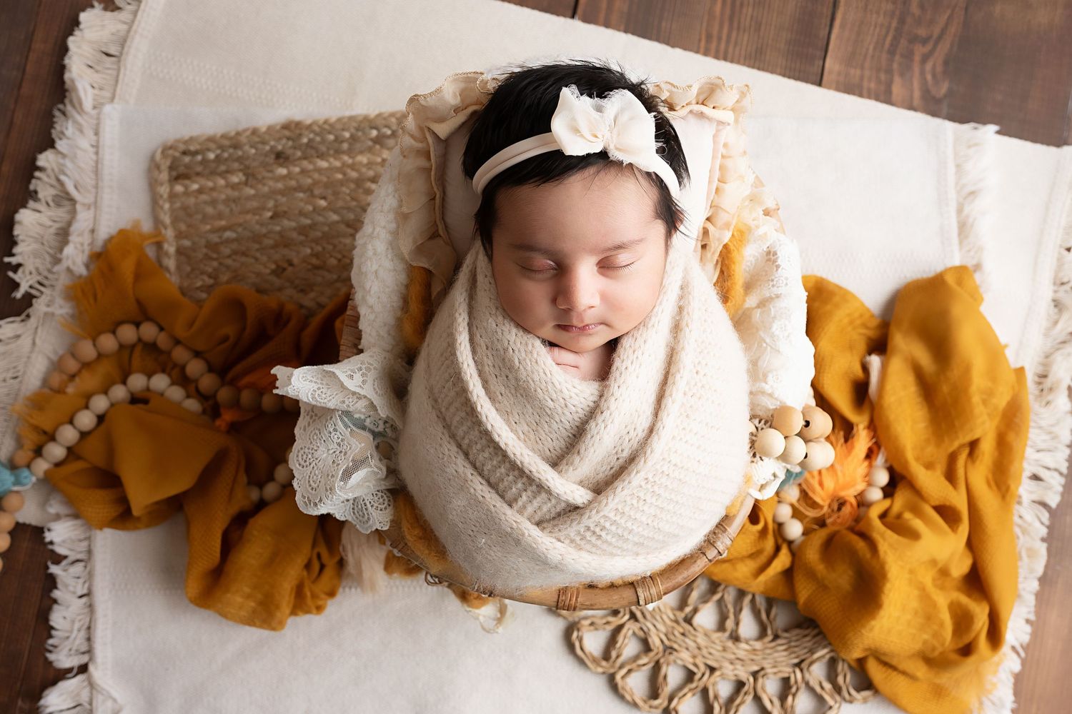 Sleeping newborn swaddled in white wrap with bow headband on cream blanket with golden accents.