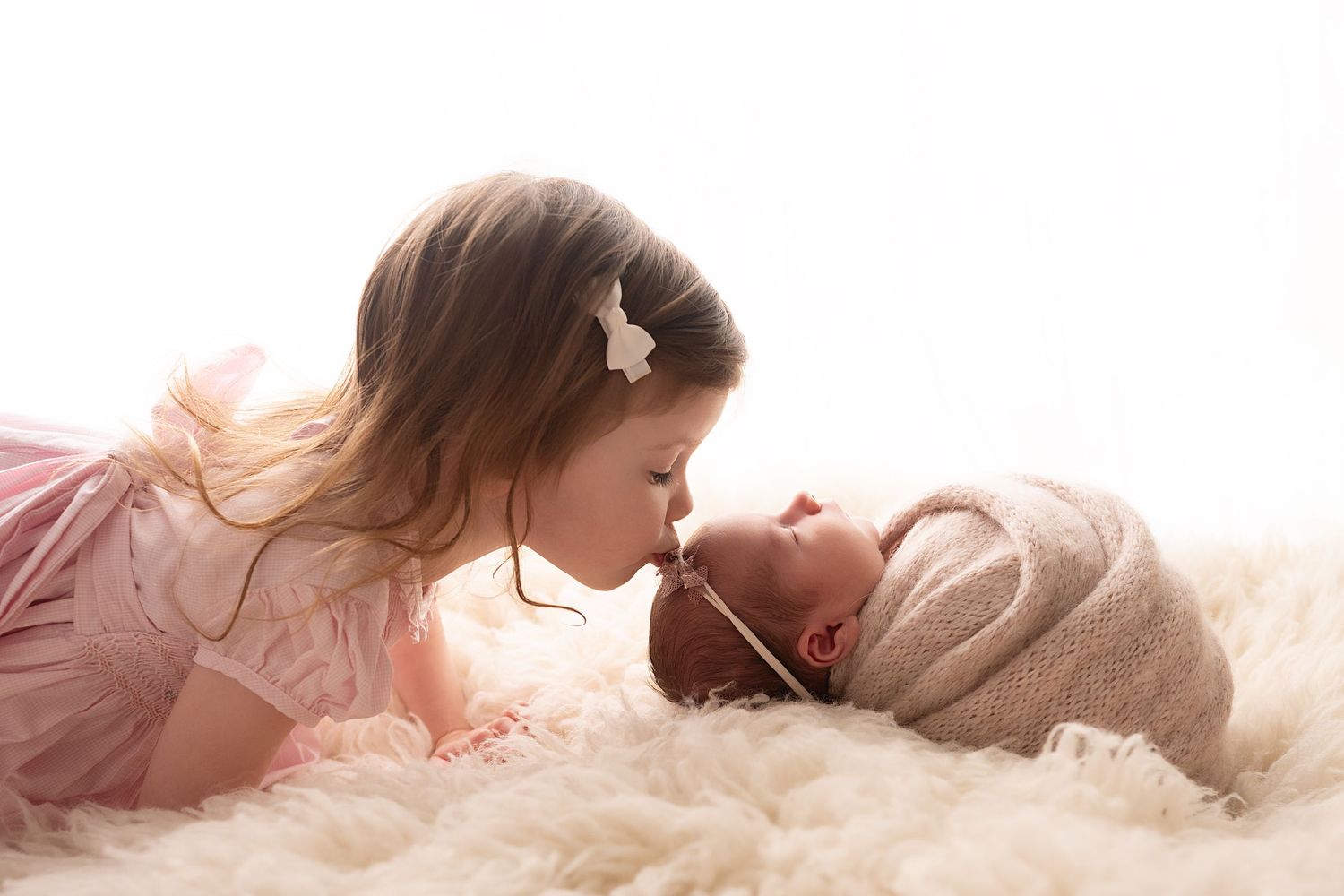 Sweet sibling moment as sister kisses newborn on fluffy rug.