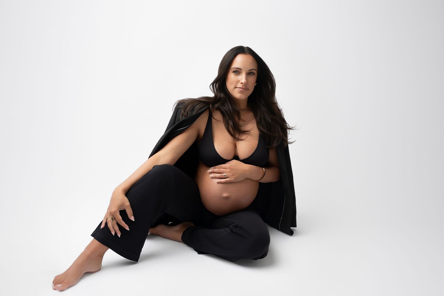 Elegant maternity portrait of woman in black outfit against white backdrop.