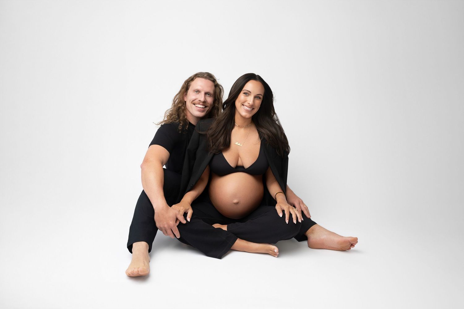 Maternity photoshoot with couple in black outfits against white backdrop.
