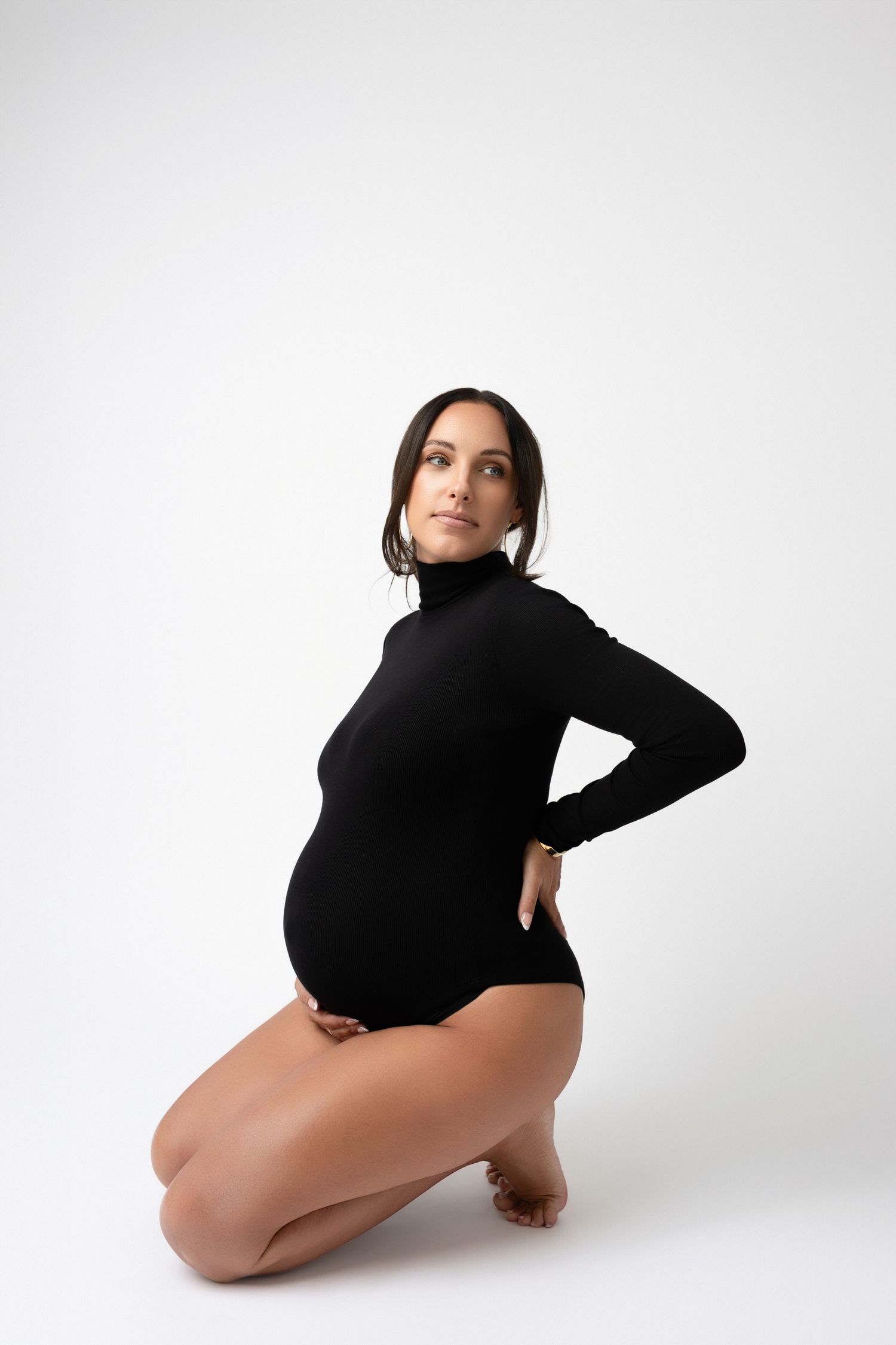 Minimalist maternity portrait of woman in black turtleneck bodysuit on white background.