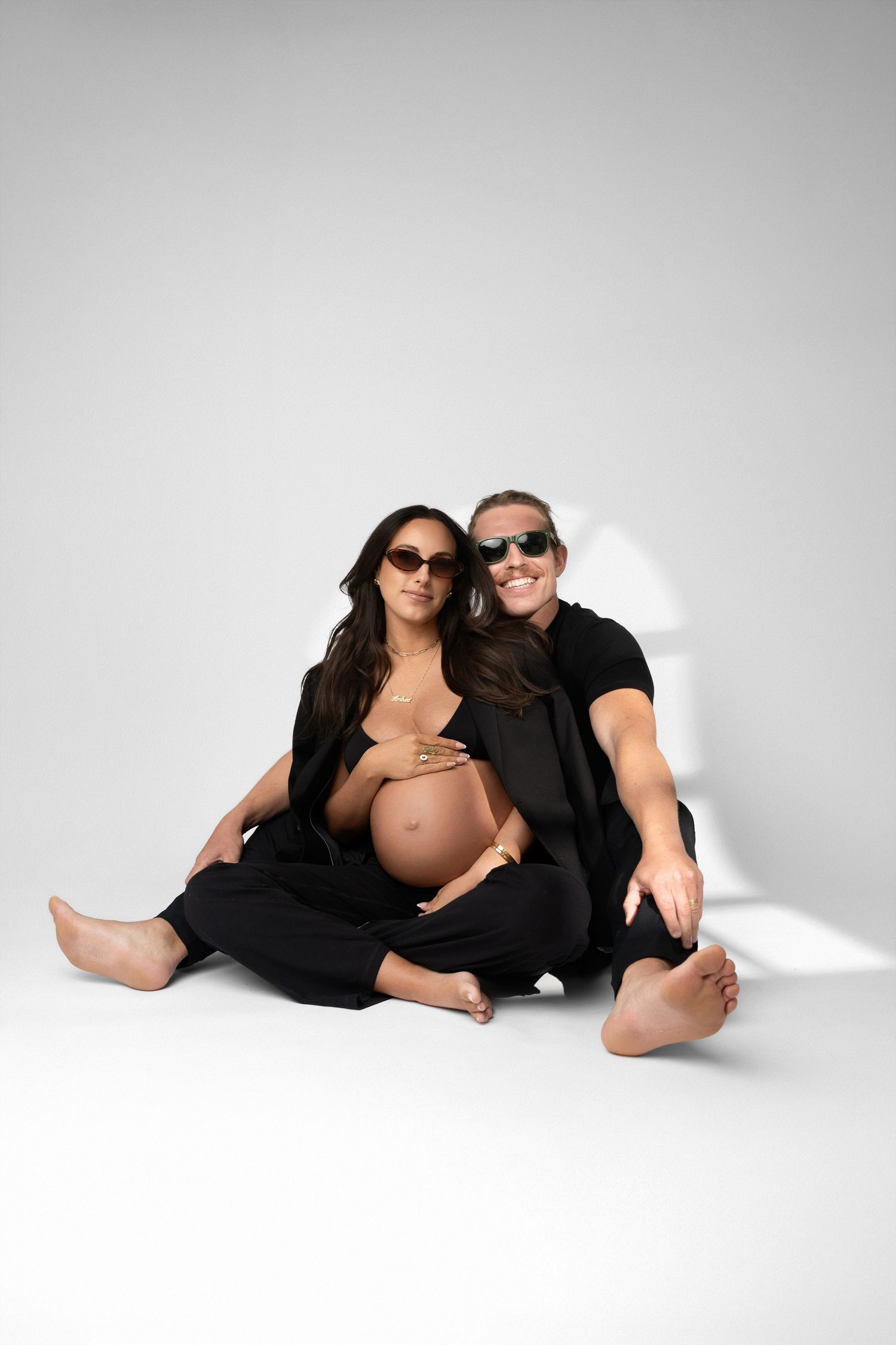 Maternity couple wearing sunglasses and black outfits posing against white background.