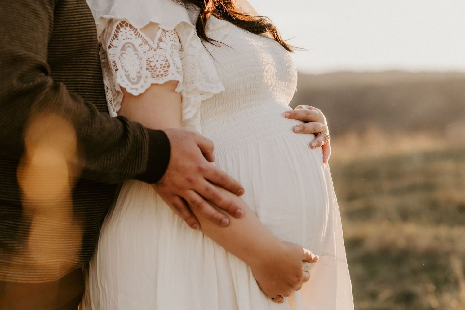 Sunlit maternity photo showing hands embracing pregnant belly in white lace dress against natural backdrop.