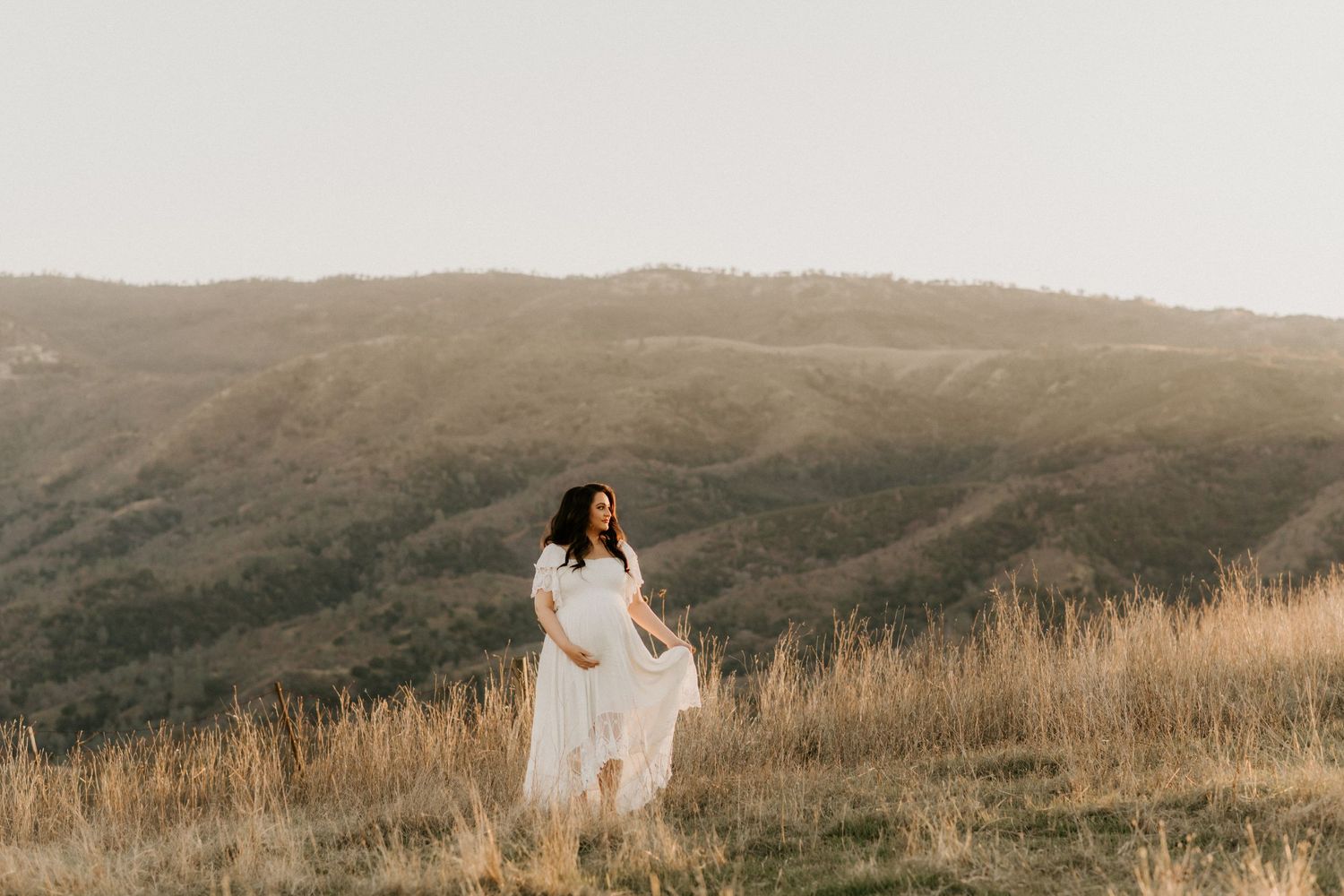 Silhouetted maternity figure in white dress standing on hilltop during golden hour.