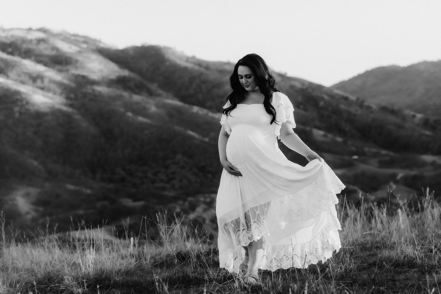 Expectant person in flowing white dress standing in mountain landscape at sunset.