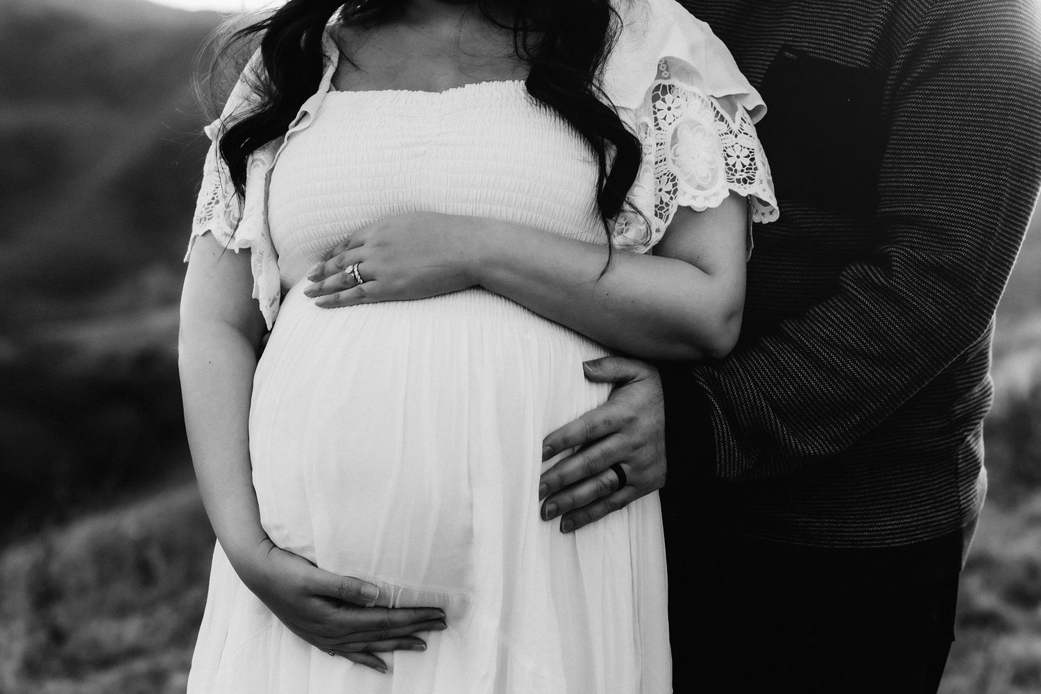 Intimate black and white maternity photo of hands cradling a pregnant belly in a white lace dress.
