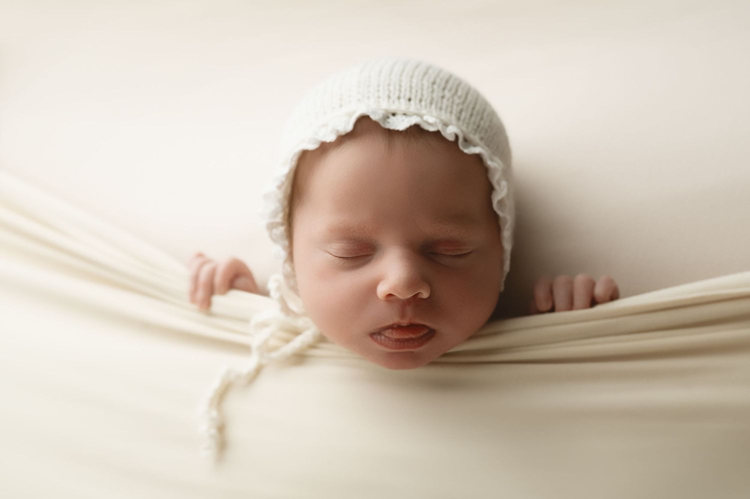Peaceful newborn wearing white bonnet nestled in cream colored wrap.