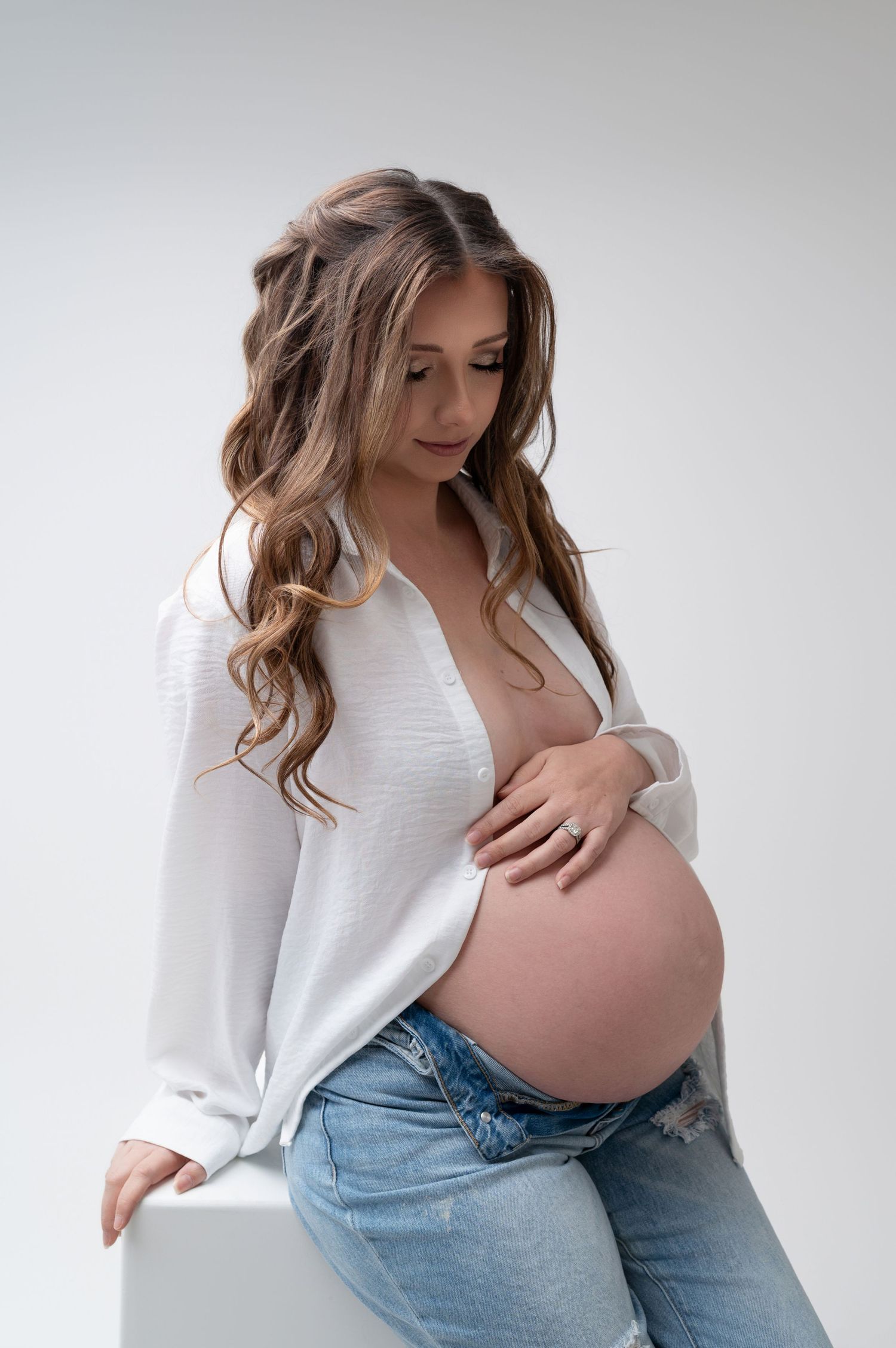 Maternity portraits showing pregnant woman in white top and jeans against light background.