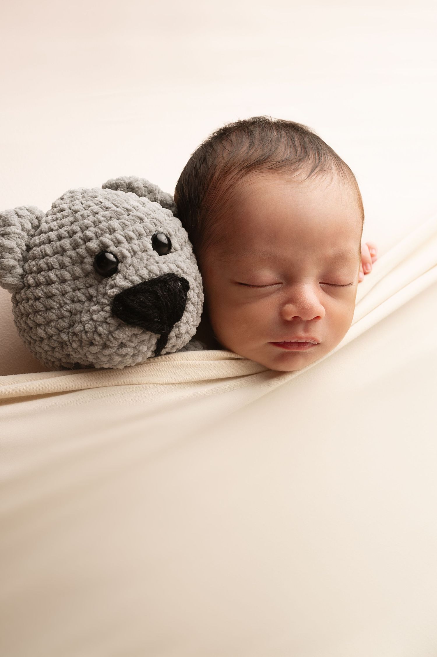 Sleeping newborn baby posed with grey knitted teddy bear on cream background.