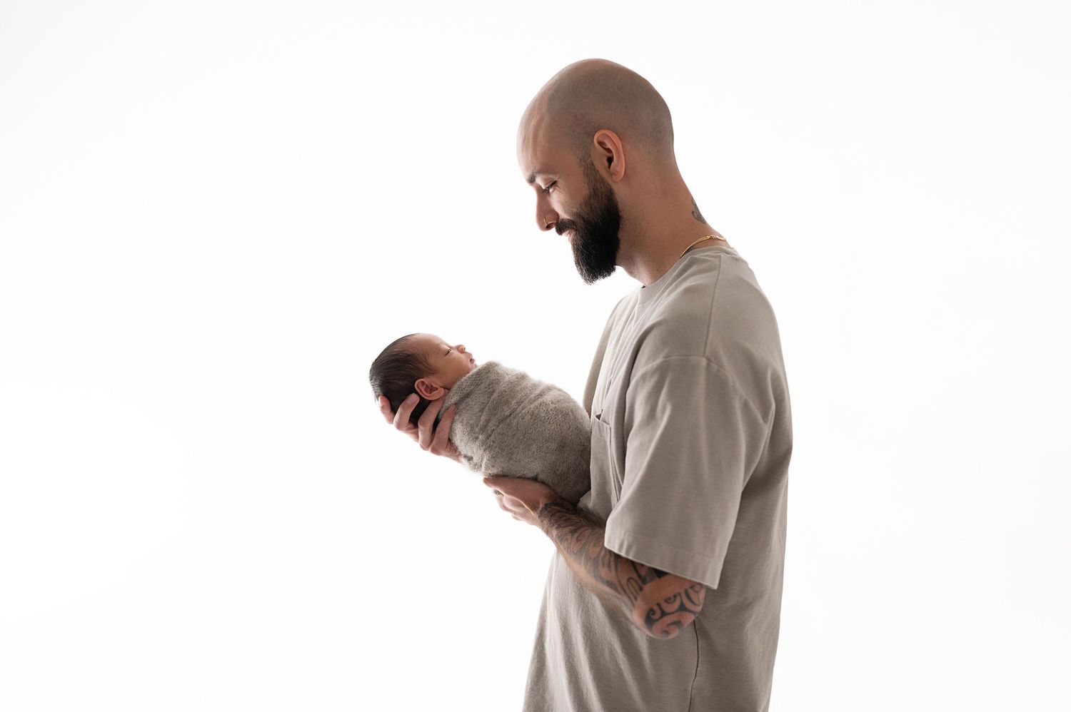 Person wearing grey shirt tenderly holds sleeping newborn baby in minimalist studio setting.