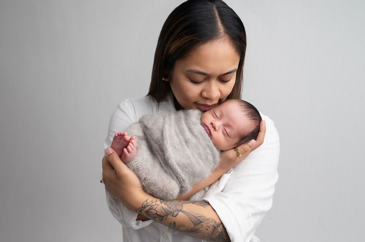 Mother in white shirt lovingly embraces sleeping newborn baby in studio portrait.