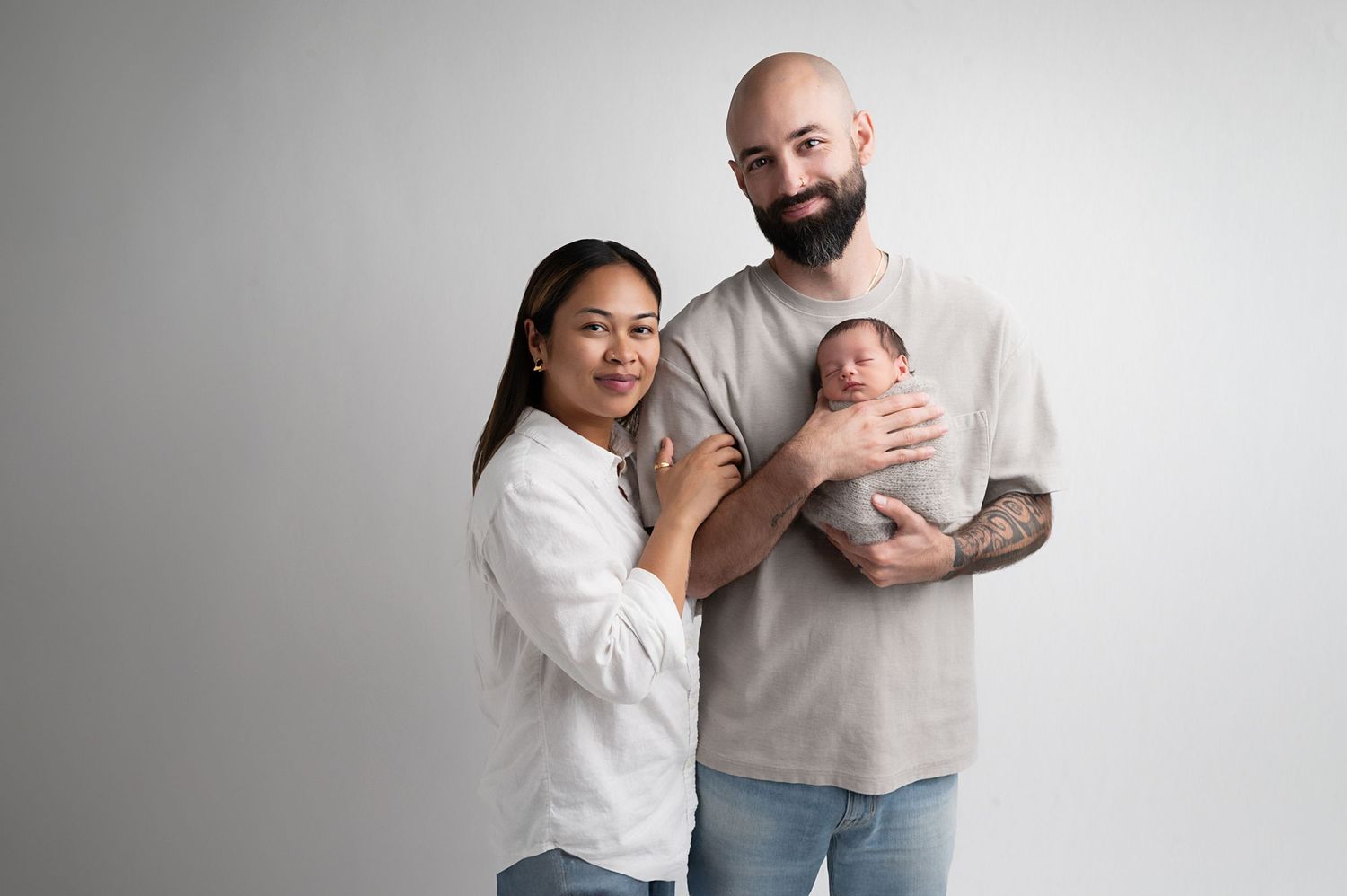 Family of three poses together with newborn baby against white studio background.