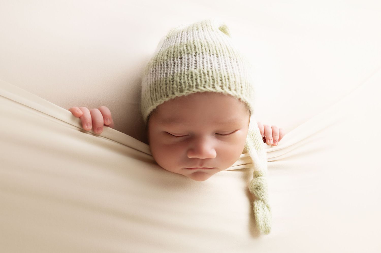 Peaceful sleeping newborn wearing knit hat photographed on cream backdrop.