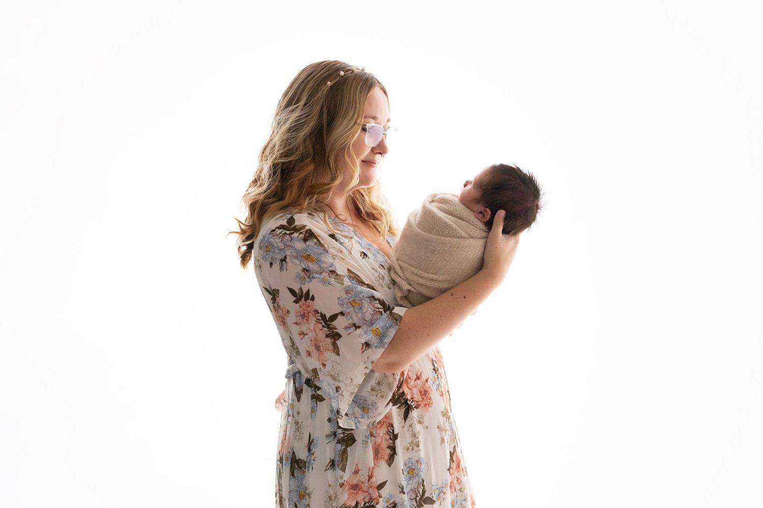 Woman in floral dress holds newborn baby against white background during portrait session.