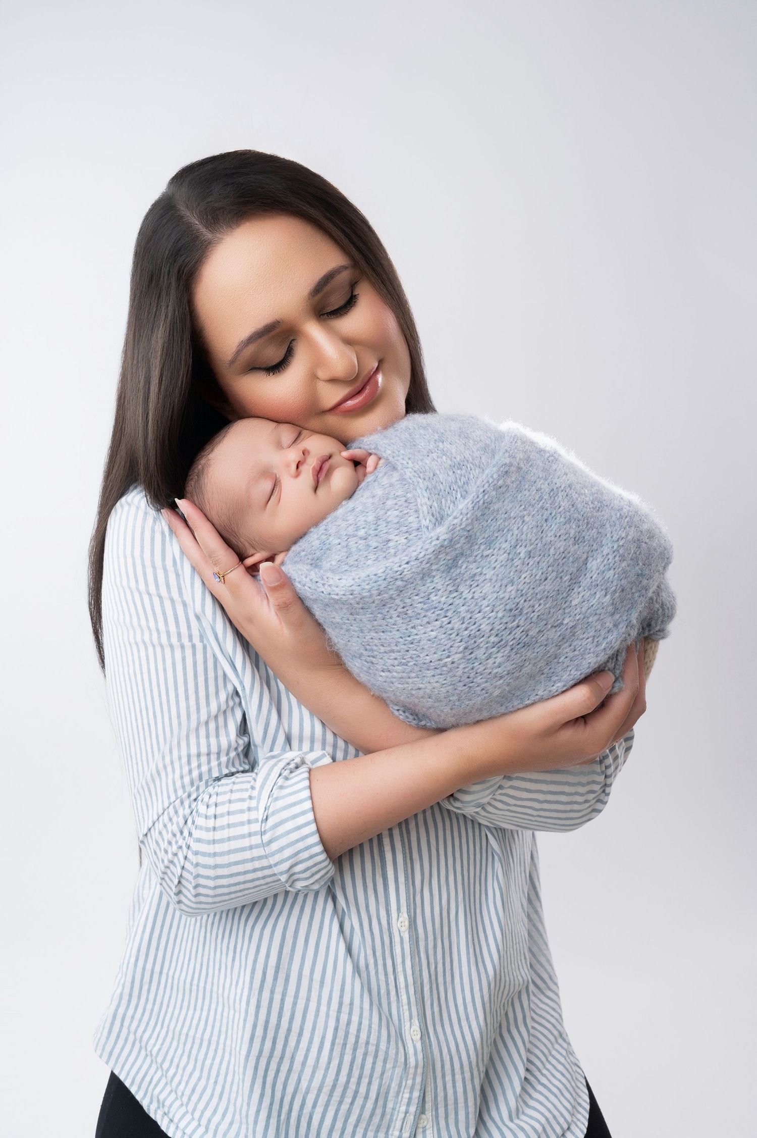 Mother in striped shirt holding newborn wrapped in gray blanket during studio photoshoot.