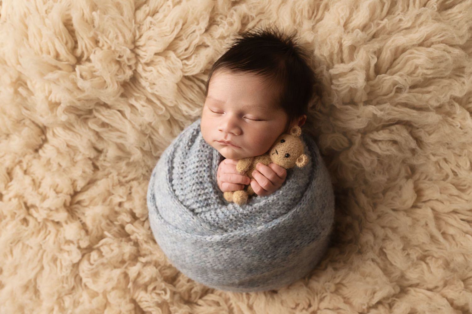 Newborn baby peacefully sleeping while wrapped in gray knit blanket on cream-colored furry rug.