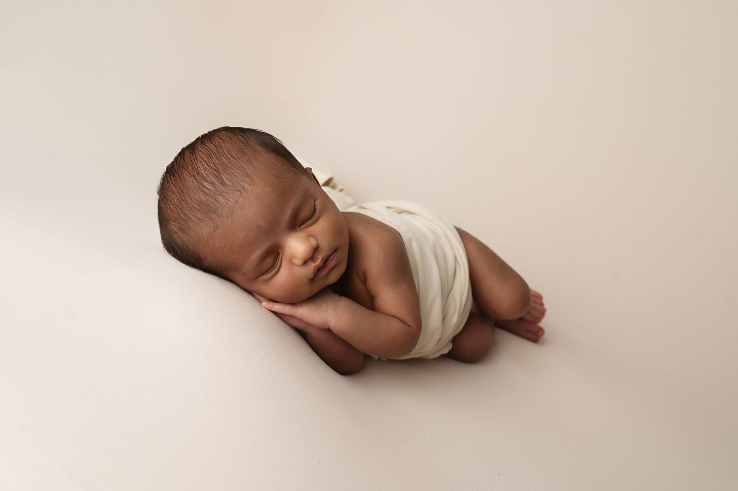 Peaceful sleeping newborn baby wearing white wrap on beige backdrop in professional studio setting.