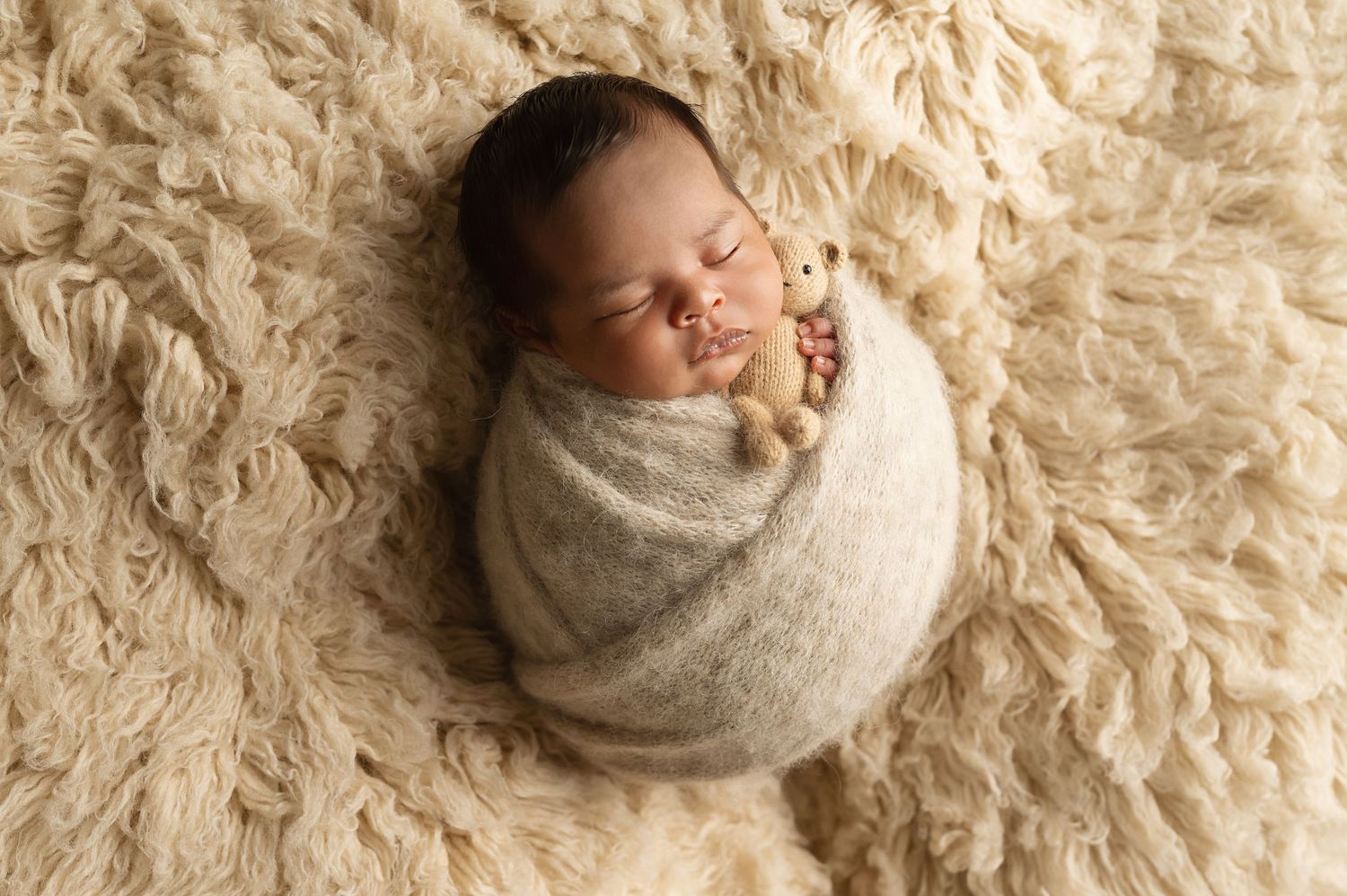 A peaceful newborn sleeps while wrapped in beige swaddling on textured cream backdrop.