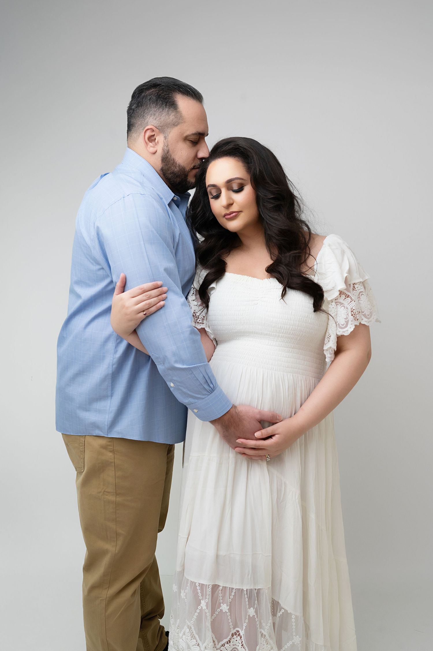 Studio maternity portrait showing couple in white dress and blue shirt against grey background.
