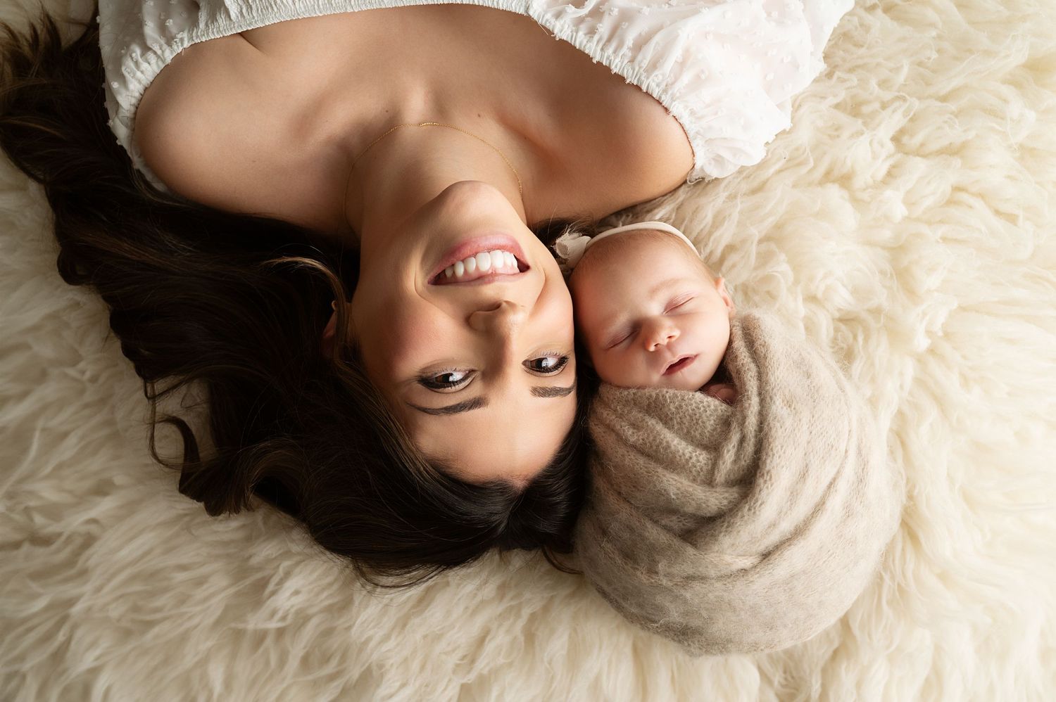 Overhead view of parent and swaddled baby lying on white fur backdrop.
