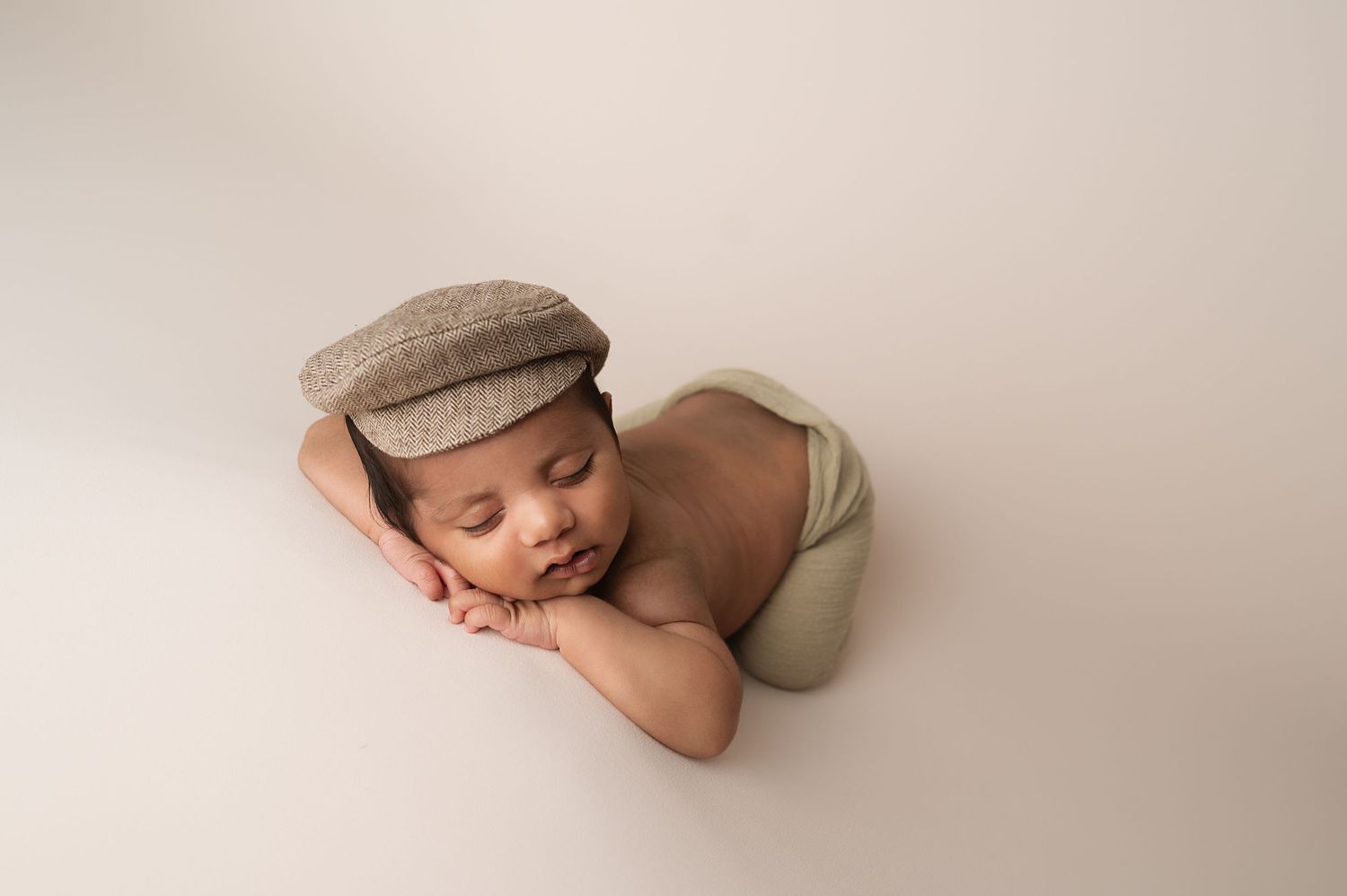 Sleeping newborn wearing a flat cap rests peacefully on a light beige background.