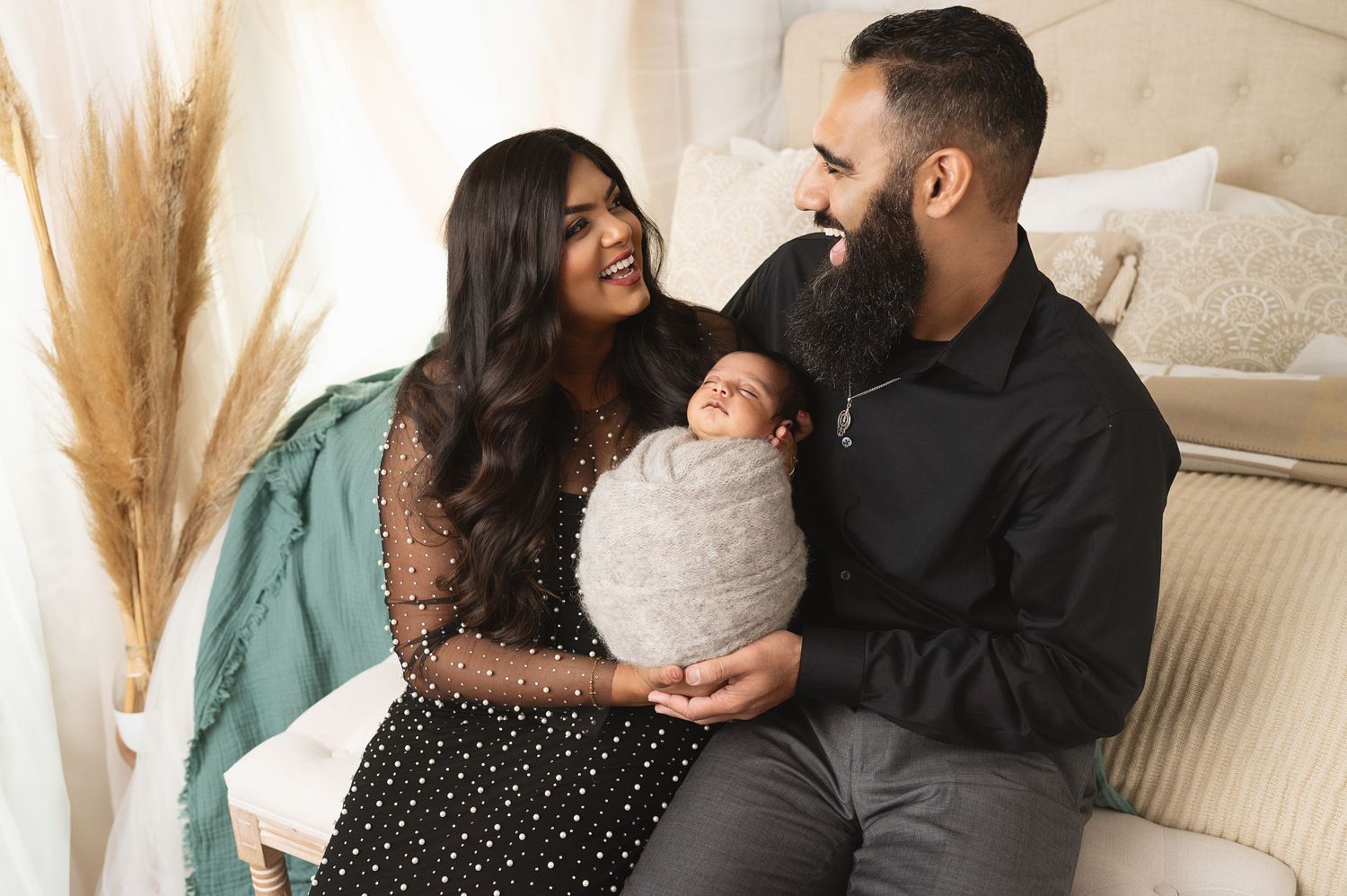 A couple sits on couch with their swaddled newborn near decorative pampas grass.